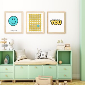 Puede incluir: Tres impresiones enmarcadas con diseños de caras sonrientes. La primera presenta una cara sonriente azul, la segunda tiene caras sonrientes amarillas con una azul, y la tercera dice "YOU". Las impresiones están sobre un mueble de almacenamiento verde menta.
