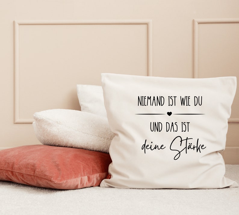 K&ouml;nnte beinhalten: Ein wei&szlig;es Kissen mit schwarzem Text, der "Niemand ist wie du und das ist deine St&auml;rke" lautet, was &uuml;bersetzt "Niemand ist wie du und das ist deine St&auml;rke" bedeutet.