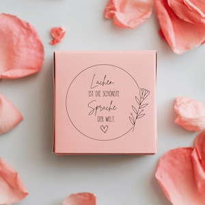 May include: A pink box with a white circle and black text that reads "Lachen ist die sch&ouml;nste Sprache der Welt."  The text is surrounded by a small flower illustration.