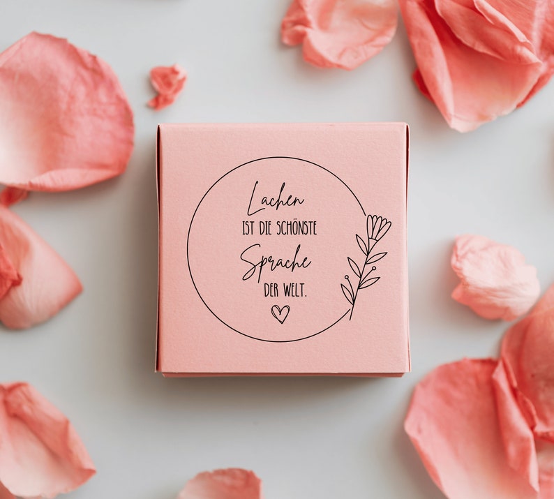 May include: A pink box with a white circle and black text that reads "Lachen ist die sch&ouml;nste Sprache der Welt."  The text is surrounded by a small flower illustration.