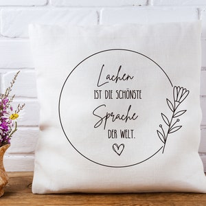 May include: White throw pillow with black text that reads "Lachen ist die sch&ouml;nste Sprache der Welt."  The text is inside a circle with a small heart at the bottom. A simple black floral design is on the right side of the pillow.