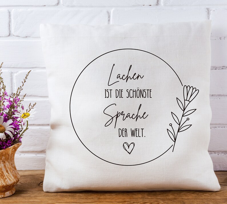 May include: White throw pillow with black text that reads "Lachen ist die sch&ouml;nste Sprache der Welt."  The text is inside a circle with a small heart at the bottom. A simple black floral design is on the right side of the pillow.