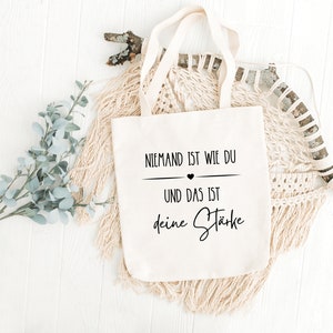 K&ouml;nnte beinhalten: Eine wei&szlig;e Stofftasche mit schwarzem Text, der "Niemand ist wie du und das ist deine St&auml;rke" lautet, was &uuml;bersetzt "Niemand ist wie du und das ist deine St&auml;rke" bedeutet.