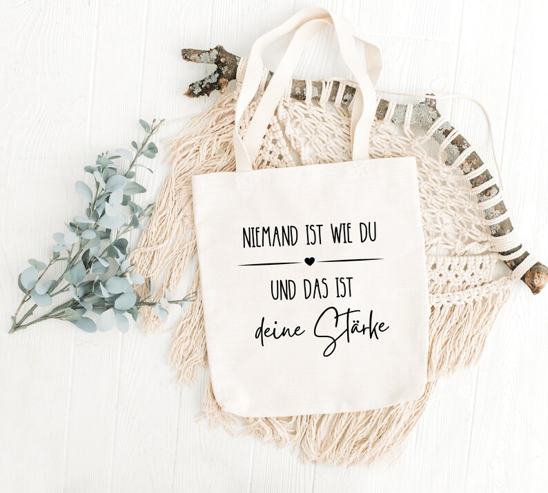 K&ouml;nnte beinhalten: Eine wei&szlig;e Stofftasche mit schwarzem Text, der "Niemand ist wie du und das ist deine St&auml;rke" lautet, was &uuml;bersetzt "Niemand ist wie du und das ist deine St&auml;rke" bedeutet.