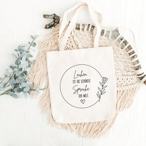 May include: A white canvas tote bag with a black circle design that says "Lachen ist die sch&ouml;nste Sprache der Welt."  The design also includes a small heart and a stylized flower.