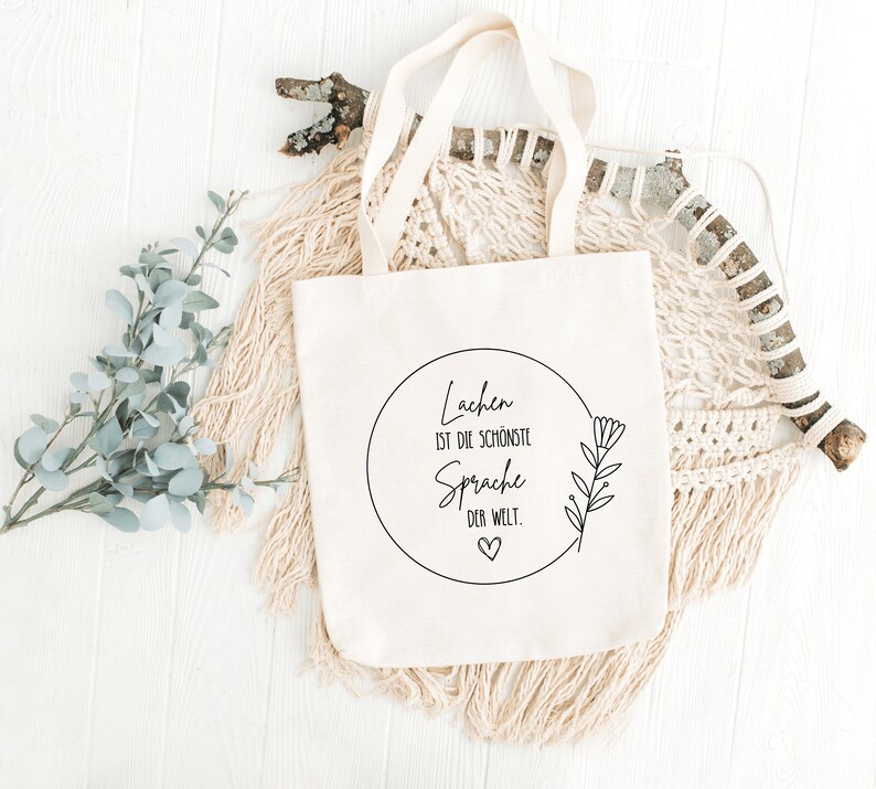 May include: A white canvas tote bag with a black circle design that says "Lachen ist die sch&ouml;nste Sprache der Welt."  The design also includes a small heart and a stylized flower.