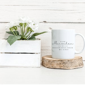 May include: White ceramic mug with black text that reads "Kleine Menschen brauchen grosse Herzen wie deines!" The mug is sitting on a wooden coaster.
