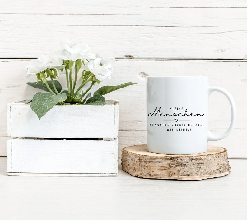 May include: White ceramic mug with black text that reads "Kleine Menschen brauchen grosse Herzen wie deines!" The mug is sitting on a wooden coaster.