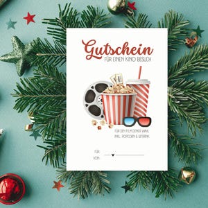 Puede incluir: Un vale de regalo blanco con el texto alemán "Gutschein für einen Kino Besuch" presenta una bobina de película, palomitas de maíz, una bebida y gafas 3D. El certificado está rodeado de decoraciones navideñas, incluyendo ramas de pino, estrellas y adornos.