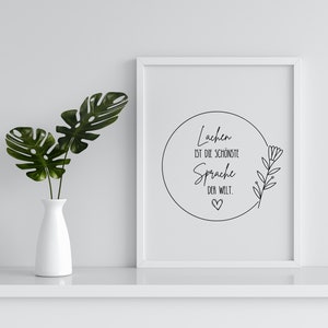 May include: A white framed print with a black circle and text that reads "Lachen ist die sch&ouml;nste Sprache der Welt."  The circle has a small heart at the bottom and a simple line drawing of a flower branch on the right side.