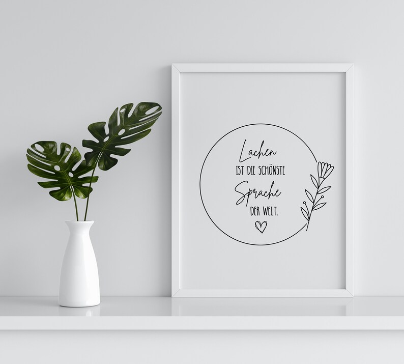 May include: A white framed print with a black circle and text that reads "Lachen ist die sch&ouml;nste Sprache der Welt."  The circle has a small heart at the bottom and a simple line drawing of a flower branch on the right side.