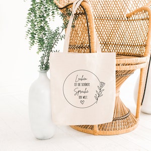 May include: A white canvas tote bag with black text that reads "Lachen ist die schoenste Sprache der Welt."  The text is in a circle with a small heart at the bottom.  The bag is sitting on a wicker chair.