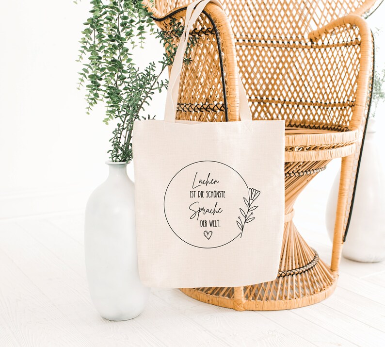May include: A white canvas tote bag with black text that reads "Lachen ist die schoenste Sprache der Welt."  The text is in a circle with a small heart at the bottom.  The bag is sitting on a wicker chair.