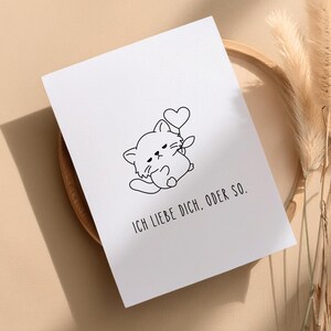 Könnte beinhalten: Weiße Grußkarte mit einer schwarzen Strichzeichnung einer Katze, die ein Herz hält. Der Text "ICH LIEBE DICH, ODER SO." ist in Schwarz unter der Katzenillustration gedruckt. Die Karte liegt auf einem Holztablett mit getrockneten Pflanzen im Hintergrund.