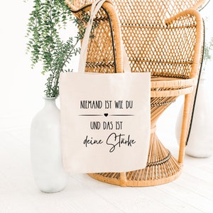 K&ouml;nnte beinhalten: Eine wei&szlig;e Stofftasche mit schwarzem Text, der "Niemand ist wie du und das ist deine St&auml;rke" lautet, was &uuml;bersetzt "Niemand ist wie du und das ist deine St&auml;rke" bedeutet.