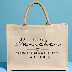 May include: A beige jute tote bag with black text that reads "Kleine Menschen brauchen grosse Herzen wie deines!"  The text is in a cursive font with a small heart symbol between the two lines of text.