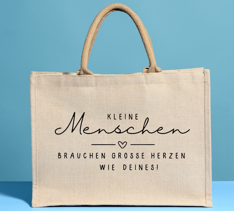 May include: A beige jute tote bag with black text that reads "Kleine Menschen brauchen grosse Herzen wie deines!"  The text is in a cursive font with a small heart symbol between the two lines of text.