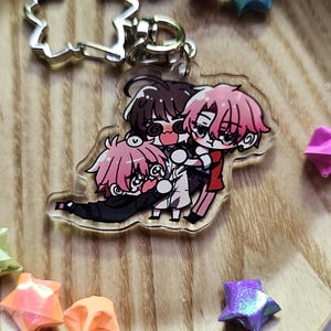 May include: A clear acrylic keychain featuring a colorful anime-style illustration of three characters interacting. The keychain includes a silver-toned metal clasp shaped like a cherry blossom flower, attached to the top of the acrylic charm.