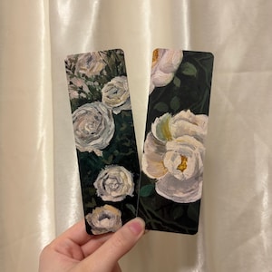 Könnte beinhalten: Zwei rechteckige Lesezeichen mit gemalten weißen Rosen auf dunkelgrünem und schwarzem Hintergrund. Das linke Lesezeichen zeigt mehrere kleine weiße Rosenblüten, während das rechte Lesezeichen eine einzelne große weiße Rosenblüte darstellt.