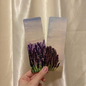 Könnte beinhalten: Zwei rechteckige Lesezeichen mit Aquarellillustrationen von Lavendelfeldern. Das linke Lesezeichen zeigt ein dichtes violettes Lavendelfeld unter einem hellen Himmel, während das rechte Lesezeichen ein kleineres Büschel dunklen Lavendels darstellt.