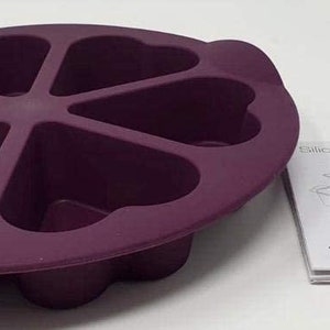 Könnte beinhalten: Lilafarbene Silikon-Backform mit sechs herzförmigen Vertiefungen. Die Form liegt auf einer weißen Oberfläche neben einem gefalteten Anleitungsblatt mit der Aufschrift "Silicone Baking Forms" und "Tupperware".