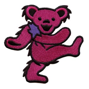 Puede incluir: Un oso rosa bailando con un parche morado en la espalda. El oso está bordado y tiene un contorno negro.