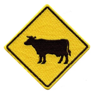 May include: Yellow diamond-shaped warning sign with a black silhouette of a cow inside. The sign is made of fabric and has a stitched border.