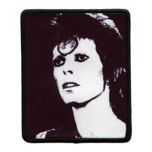 Puede incluir: Imagen en blanco y negro de David Bowie con un diseño de maquillaje en forma de estrella en la frente. La imagen está sobre un fondo de tela oscura.