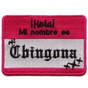 May include: A pink and white embroidered patch with the text "Hola! Mi nombre es Chingona" in black lettering. The patch has two small stars on the right side.