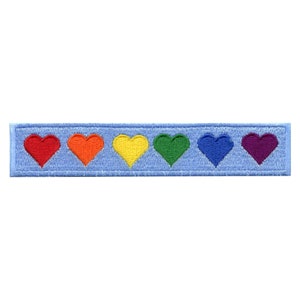 Pode incluir: Um remendo de tecido azul com seis corações bordados nas cores do arco-íris. Os corações são vermelho, laranja, amarelo, verde, azul e roxo.
