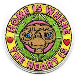 以下が含まれることがあります： E.T.の漫画の画像が描かれた黄色と緑の刺繍パッチ。画像の周りのテキストは「Home is where the heart is」で、テキストの両側にピンクのハートがあります。