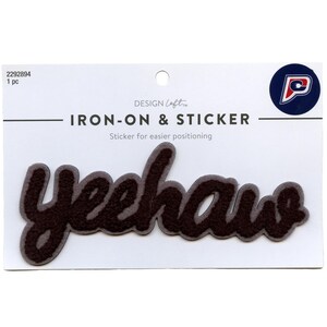 Puede incluir: Parche de plancha de fieltro marrón con la palabra "yeehaw" en una fuente cursiva. El parche está sobre un fondo blanco con el texto "DESIGN Loft IRON-ON & STICKER Sticker para un posicionamiento más fácil" arriba.