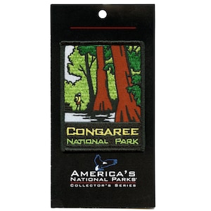 May include: A green, yellow, and brown embroidered patch with a depiction of a forest with two large trees and a person walking in the distance. The text "CONGAREE NATIONAL PARK" is printed in yellow and green on the patch.