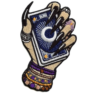 May include: An embroidered patch of a hand holding a tarot card with an eye design. The hand is adorned with bracelets and rings. The card is blue with white and gold accents.