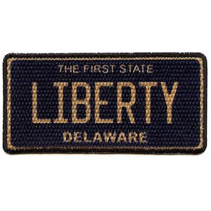 Puede incluir: Un parche rectangular con fondo azul marino y letras doradas. El texto dice "THE FIRST STATE", "LIBERTY" y "DELAWARE". El parche tiene un borde negro y una apariencia texturizada.