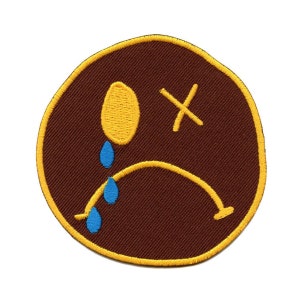 May include: A brown embroidered patch with a yellow outline. The patch depicts a sad face with a yellow eye, a blue tear, and an 'X' over the other eye.
