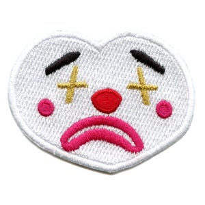 Könnte beinhalten: Weißer herzförmiger Patch mit einem traurigen Clown-Gesicht. Der Clown hat schwarze Augenbrauen, eine rote Nase und einen rosa Mund. Die Augen sind mit goldenen X durchgestrichen.