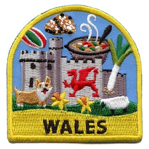 Op de afbeelding: Een kleurrijk geborduurd patch met een Welshe draak, een kasteel, een prei, een narcis, een schaap, een corgi en een kom stoofpot. De patch is geel met het woord "WALES" in zwarte letters.