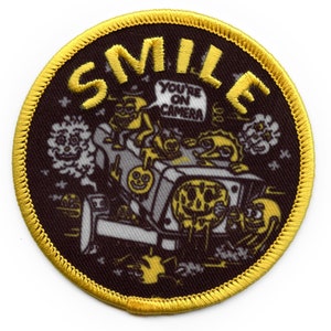 May include: A round, yellow-trimmed patch with a black background. The patch features a cartoon illustration of a vehicle with smiling faces and the word "SMILE" in yellow letters. The vehicle has a speech bubble that says "YOU'RE ON CAMERA".