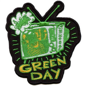 Puede incluir: Parche bordado con un televisor verde con el texto "GREEN DAY" en amarillo. El televisor tiene una pantalla verde con un gráfico de mano. El parche tiene un borde negro y un diseño floral blanco y verde.