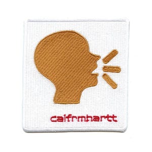 Op de afbeelding: Een witte geborduurde patch met een gouden silhouet van een persoon die spreekt met geluidgolven die uit hun mond komen. De tekst "carhartt" is onderaan de patch in rood geborduurd.