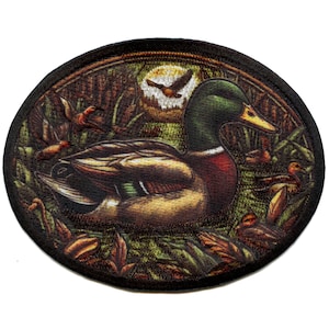 May include: An oval-shaped embroidered patch featuring a detailed image of a mallard duck in a natural setting. The duck is depicted in a realistic style with brown, green, and white feathers. The background features a lush green forest with brown leaves and a flying bird.