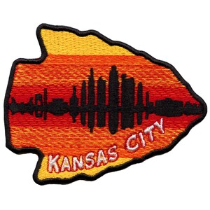 Puede incluir: Un parche bordado en forma de punta de flecha con un fondo a rayas rojo, naranja y amarillo. El parche presenta una silueta negra del horizonte de Kansas City y el texto "KANSAS CITY" en blanco.