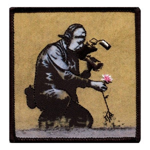 May include: A black and white embroidered patch depicting a person holding a pink flower. The person is wearing a helmet and holding a camera. The background is a tan color.