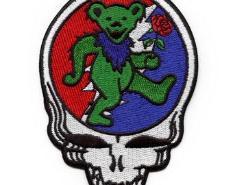 May include: A green bear with a blue collar and a red rose is walking on a red, white, and blue background inside a white skull with black eyes.