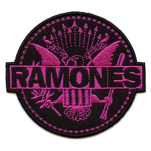 May include: A black and pink embroidered patch featuring the Ramones logo. The logo is a stylized eagle with the band's name written across the bottom.