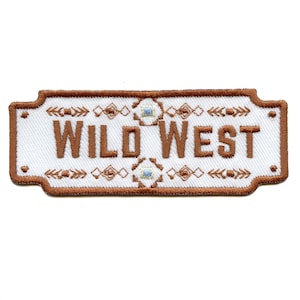 May include: A brown embroidered patch with a white background and the text "WILD WEST" in brown lettering. The patch has a decorative border with a geometric pattern.