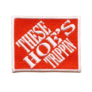 以下が含まれることがあります： オレンジと白の刺繍パッチで、"These Hoes Trippin"というテキストが書かれています