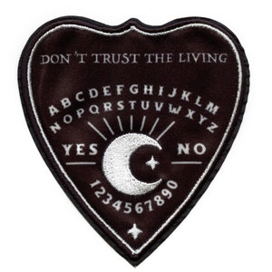 Peut inclure: Un patch en forme de cœur noir avec des coutures blanches. Le patch présente un design de planche Ouija avec les lettres A-Z, les chiffres 1-9 et les mots "YES" et "NO". Le texte "DON'T TRUST THE LIVING" est imprimé en haut du patch.