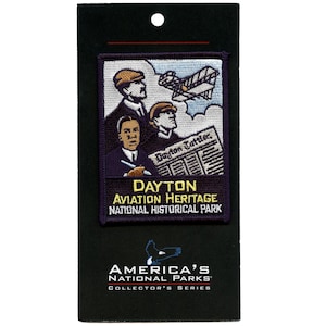 May include: A purple and yellow patch featuring an image of three men and an aeroplane. The text reads "Dayton Aviation Heritage National Historical Park" and "America's National Parks Collector's Series".